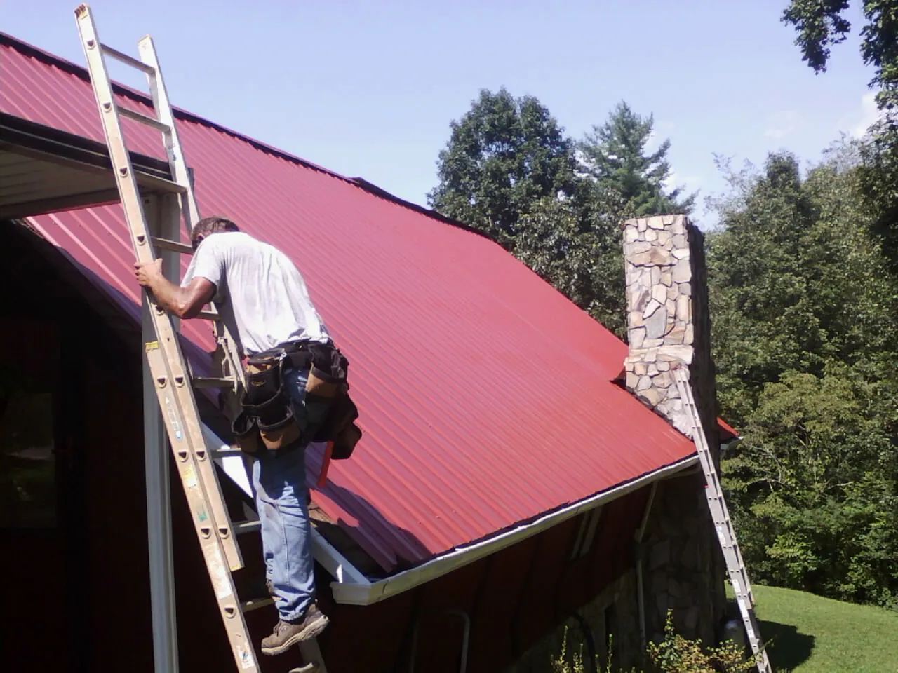 Roofer on a ladder beside a red metal roof and stone chimney — Soffit & Fascia Repair by Roofing Brothers in Odenton